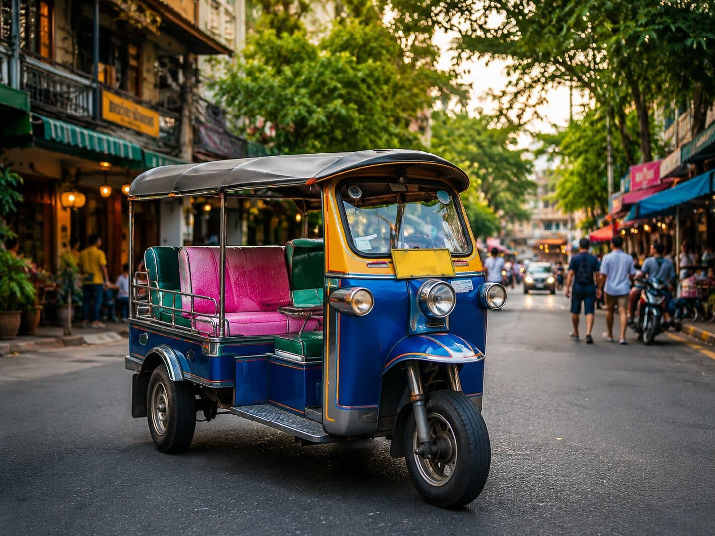 A tuk-tuk parked on a tree-lined Bangkok street with shopfronts and pedestrians.