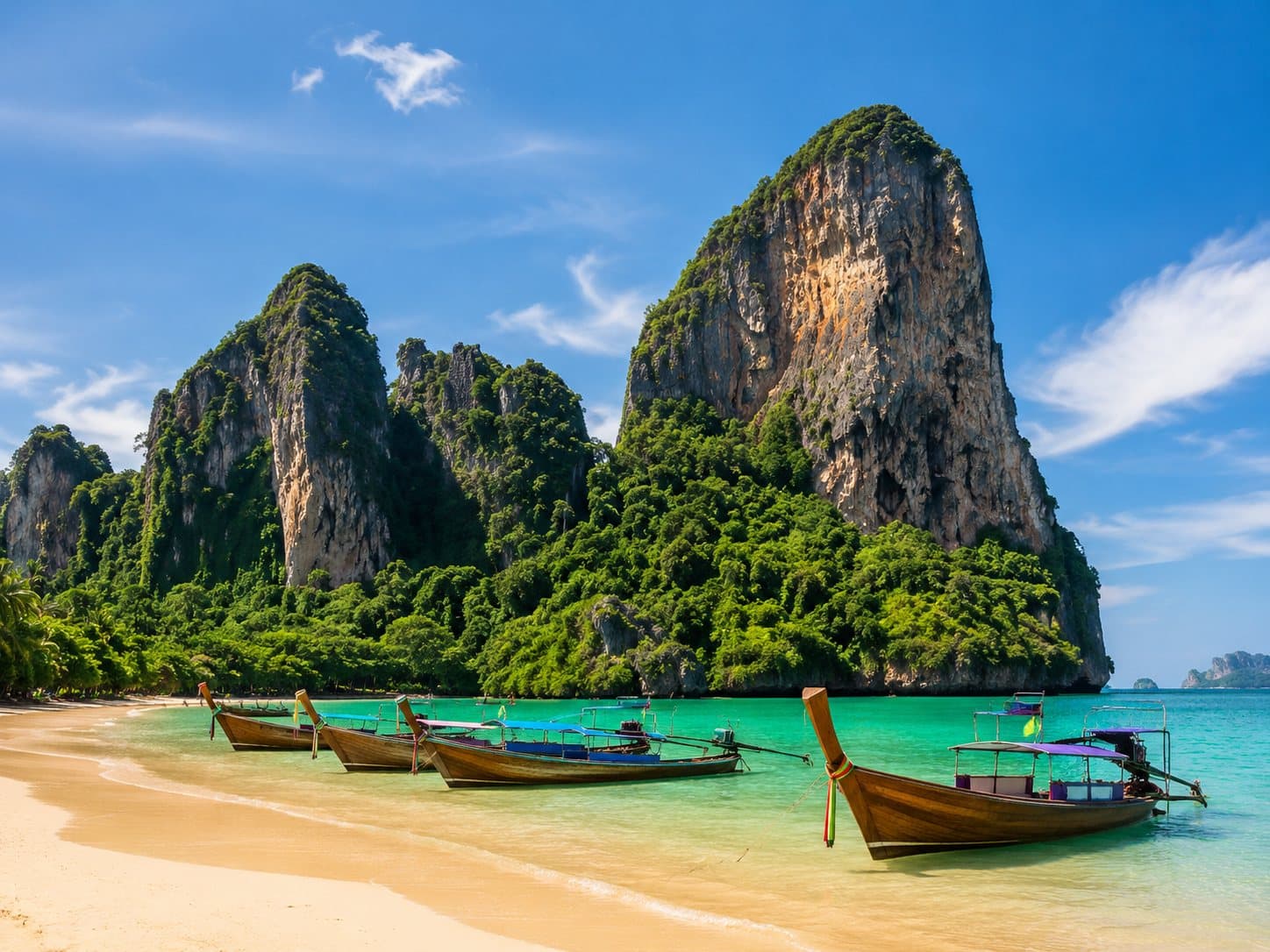 Longtail boats moored at a sandy beach below limestone cliffs on Thailand's southern coast.